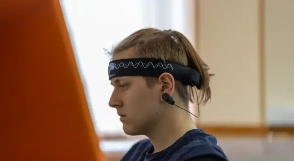 Młody mężczyzna w opasce z elektrodami do eeg w czuwaniu, skupiony na czymś poza kadrem.