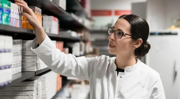 Farmaceutka w białym kitlu sięga po lek, być może pentoksyfilinę, z aptecznej półki pełnej opakowań.