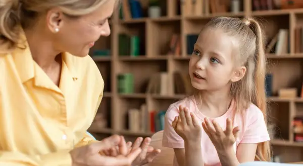 Kobieta i dziewczynka rozmawiają, gestykulując. W tle regały z książkami.
