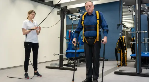 Starszy mężczyzna w specjalistycznej uprzęży i z kulami ćwiczy pod okiem fizjoterapeutki. To kluczowy etap rehabilitacji neurologicznej.