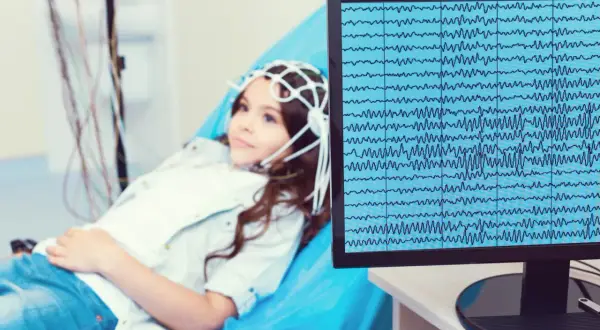 Dziewczynka z czepkiem do eeg leży na łóżku, a na monitorze widać zapis fal mózgowych.